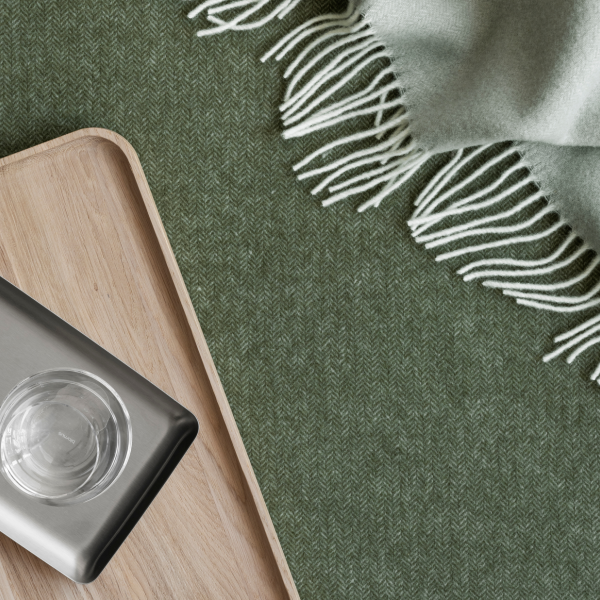 Cozy green blanket with fringes beside a wooden tray and glass on textured surface.