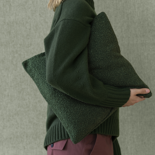 Gemütliche grüne Kissen, die von einer Person in einem stylischen Strickpullover vor einem strukturierten Hintergrund gehalten werden.