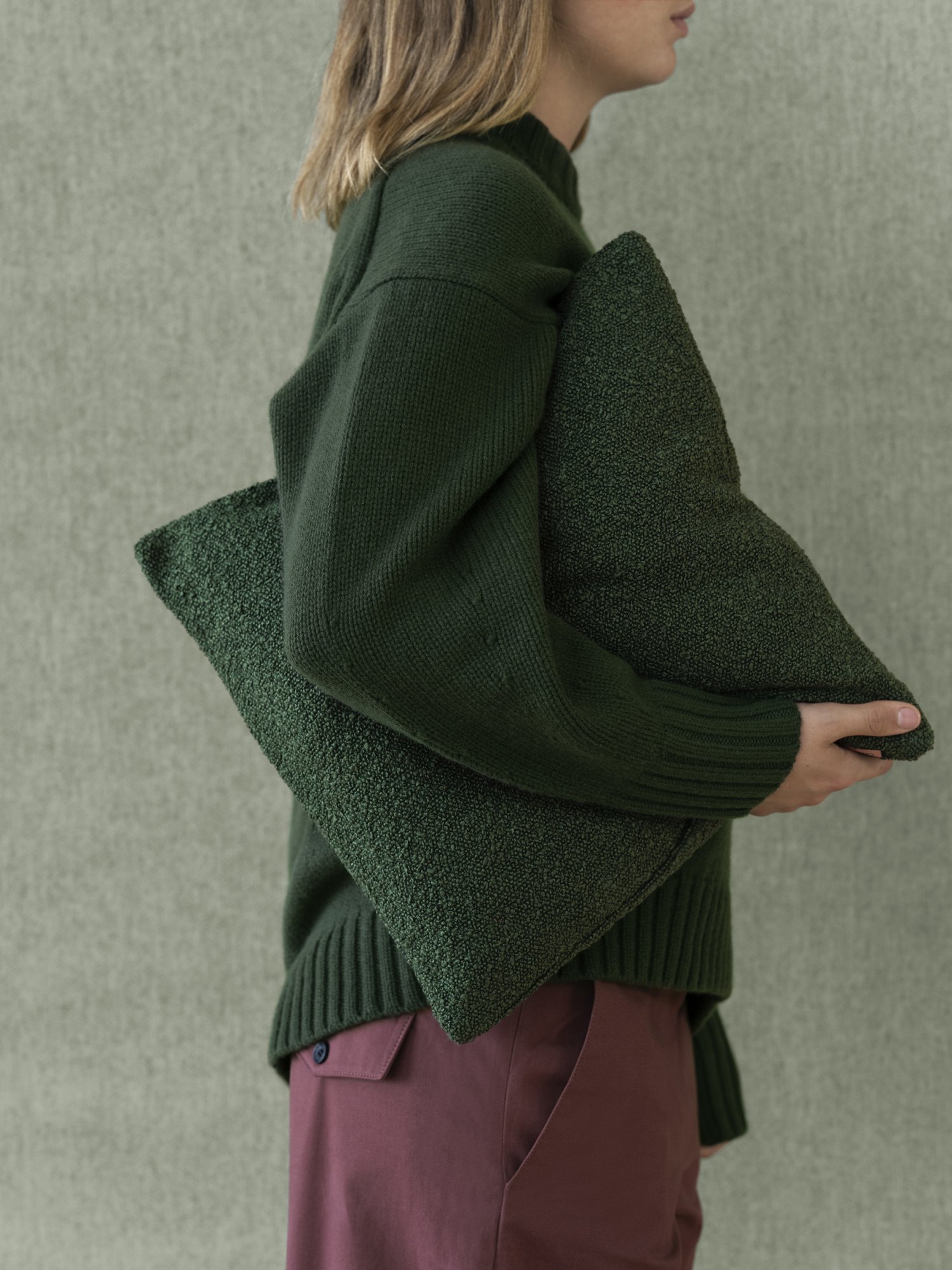 Gemütliche grüne Kissen, die von einer Person in einem stylischen Strickpullover vor einem strukturierten Hintergrund gehalten werden.